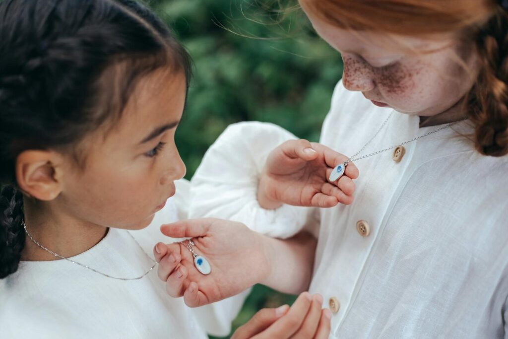 Two young girls admiring each other's necklaces in a park setting, showcasing friendship and innocence.
